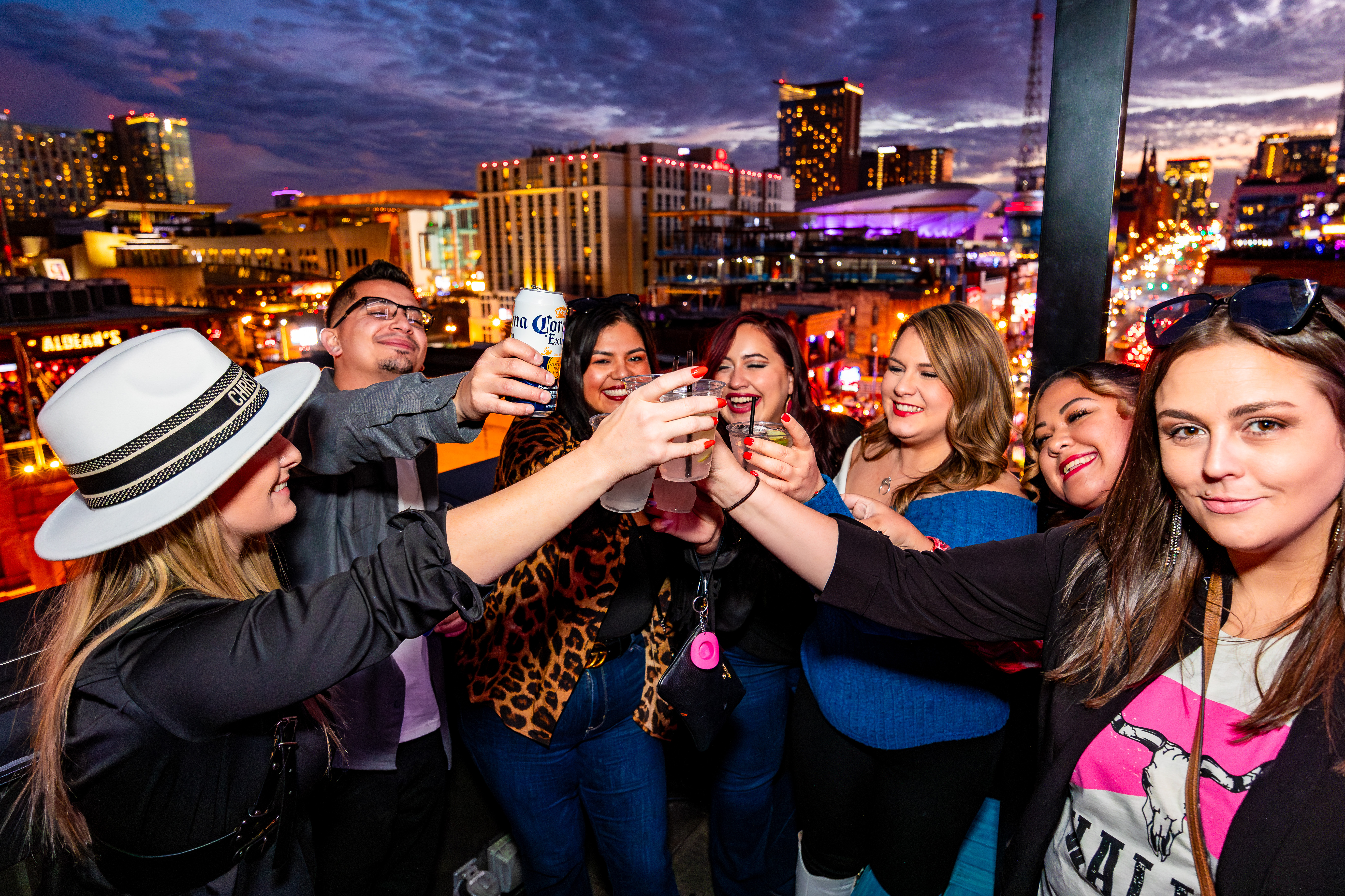 A group of friends toast their drinks on Ole Red's rooftop, with the nighttime skyline of Nashville's lower Broadway visible in the background 
