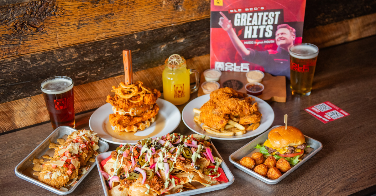 Table spread of Ole Red menu items, including nachos, bluetick burger and tots, fried green tomatoes, chicken and waffles, chicken and french fries, and an assortment of drinks.